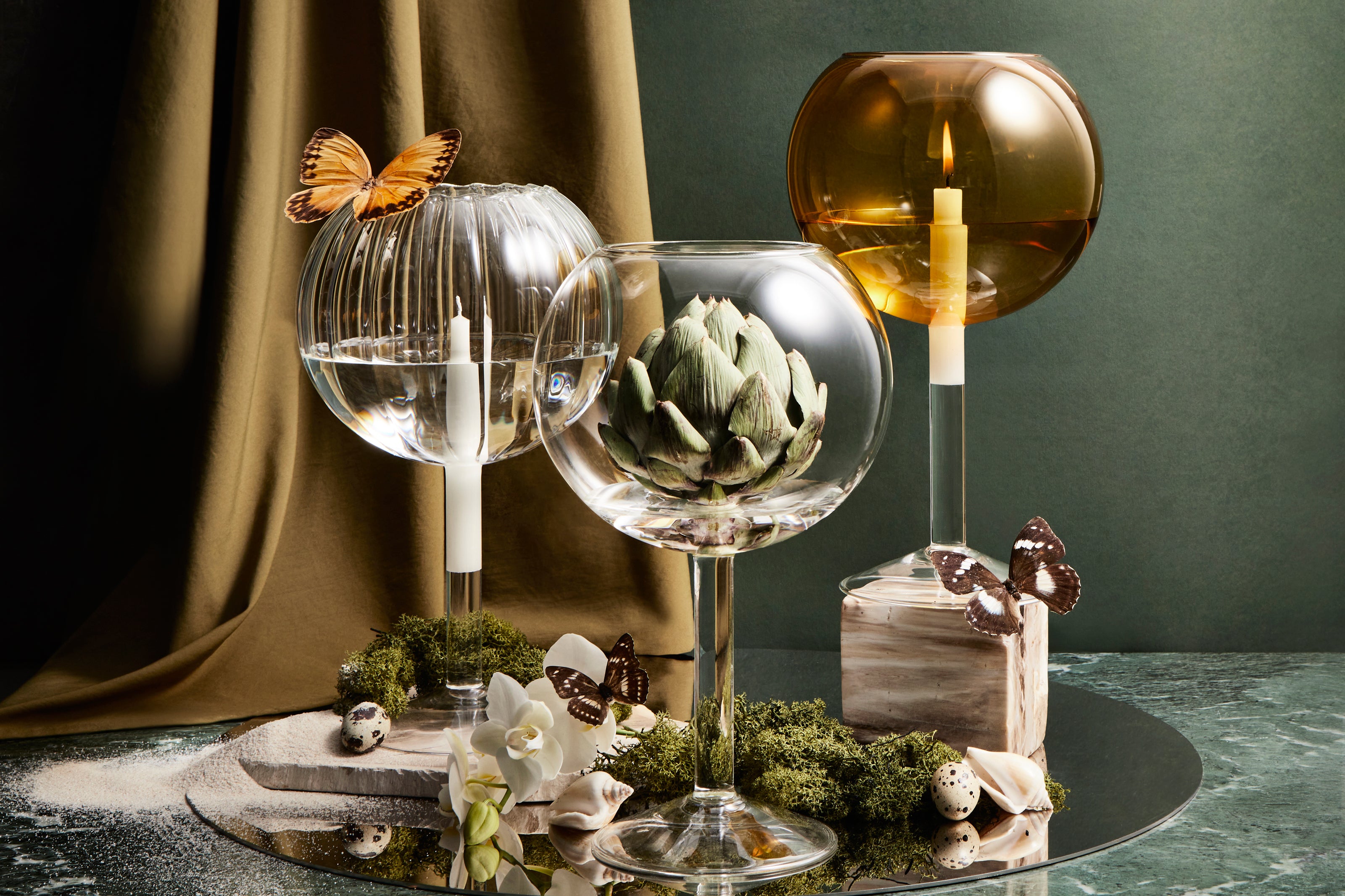 Decorative table setting with glass candleholders, dressed with butterflies, moss, an egg and an artichoke, sitting on a circular steel mirror.