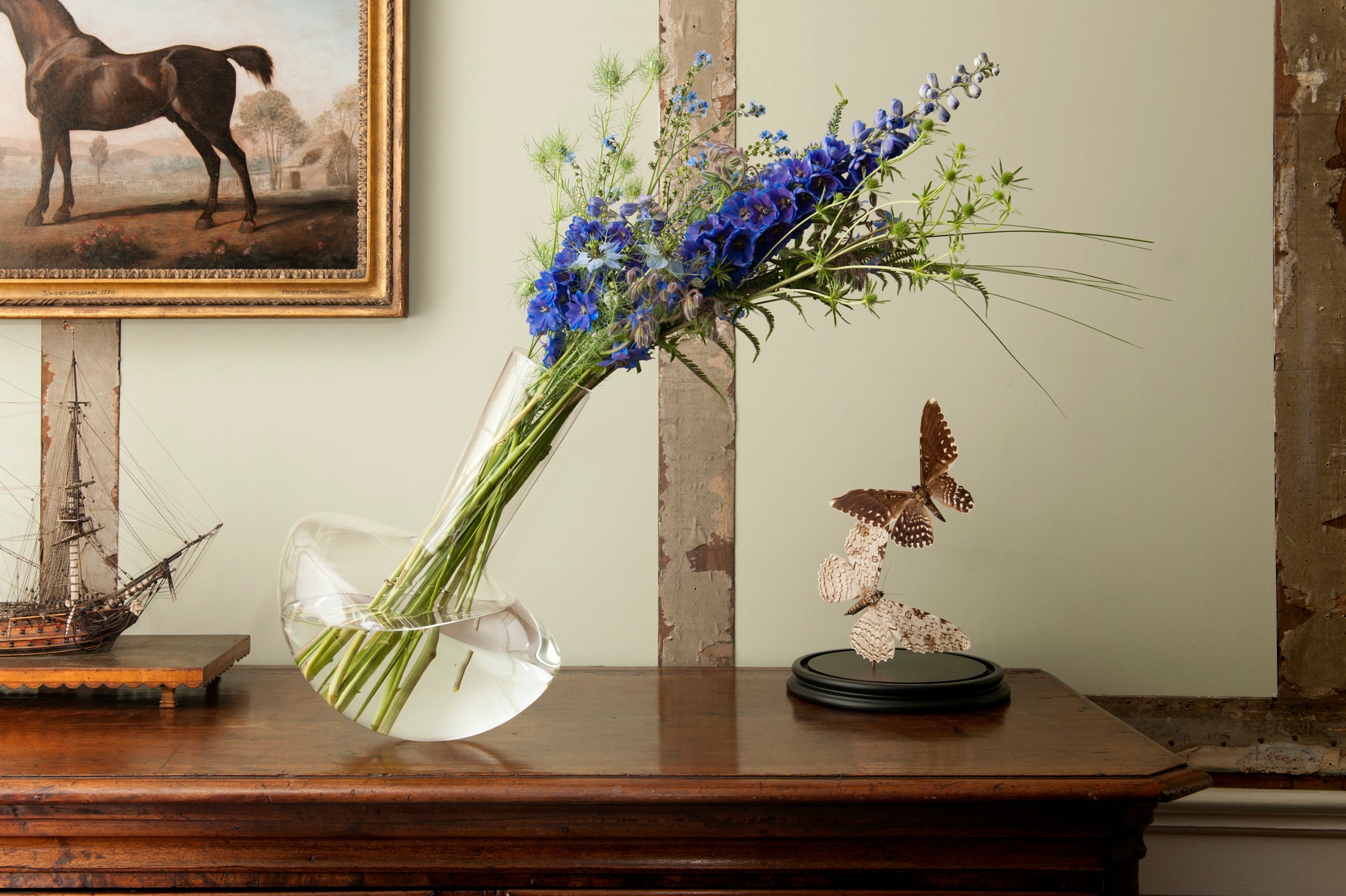 A borosilicate vase contains flowers on a wooden chest of draws. A model ship sits to its left. A framed picture of a horse hangs in the background, its head out of view.