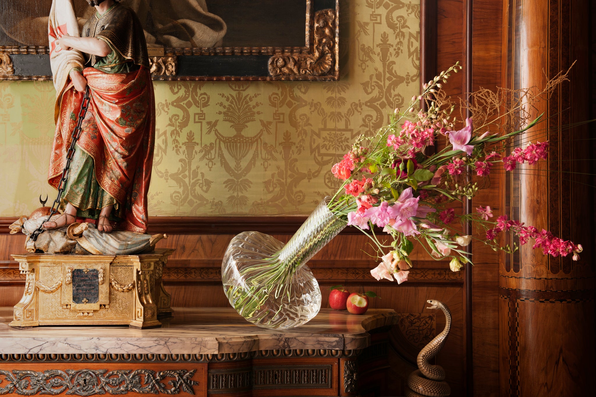 Decorative setting with a statue, vase of flowers, and apples on a wooden surface. A bite has been taken from an apple, and a snake recoils away from the vase as it leans towards it.