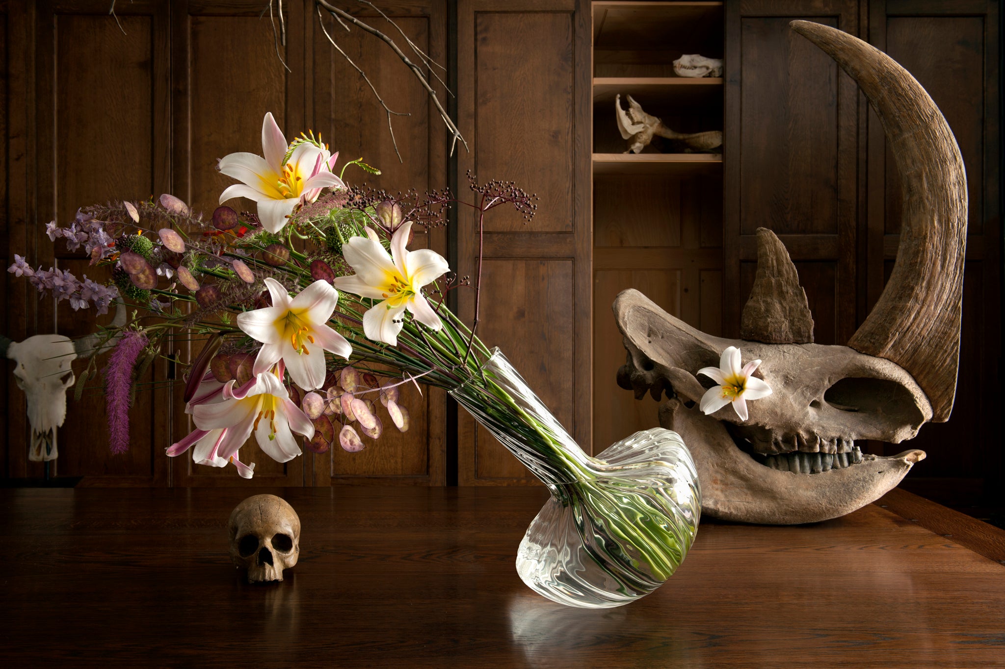 Still life arrangement with flowers, skulls, and a vase on a wooden surface. The rhino skull has a lily flower in its eye.