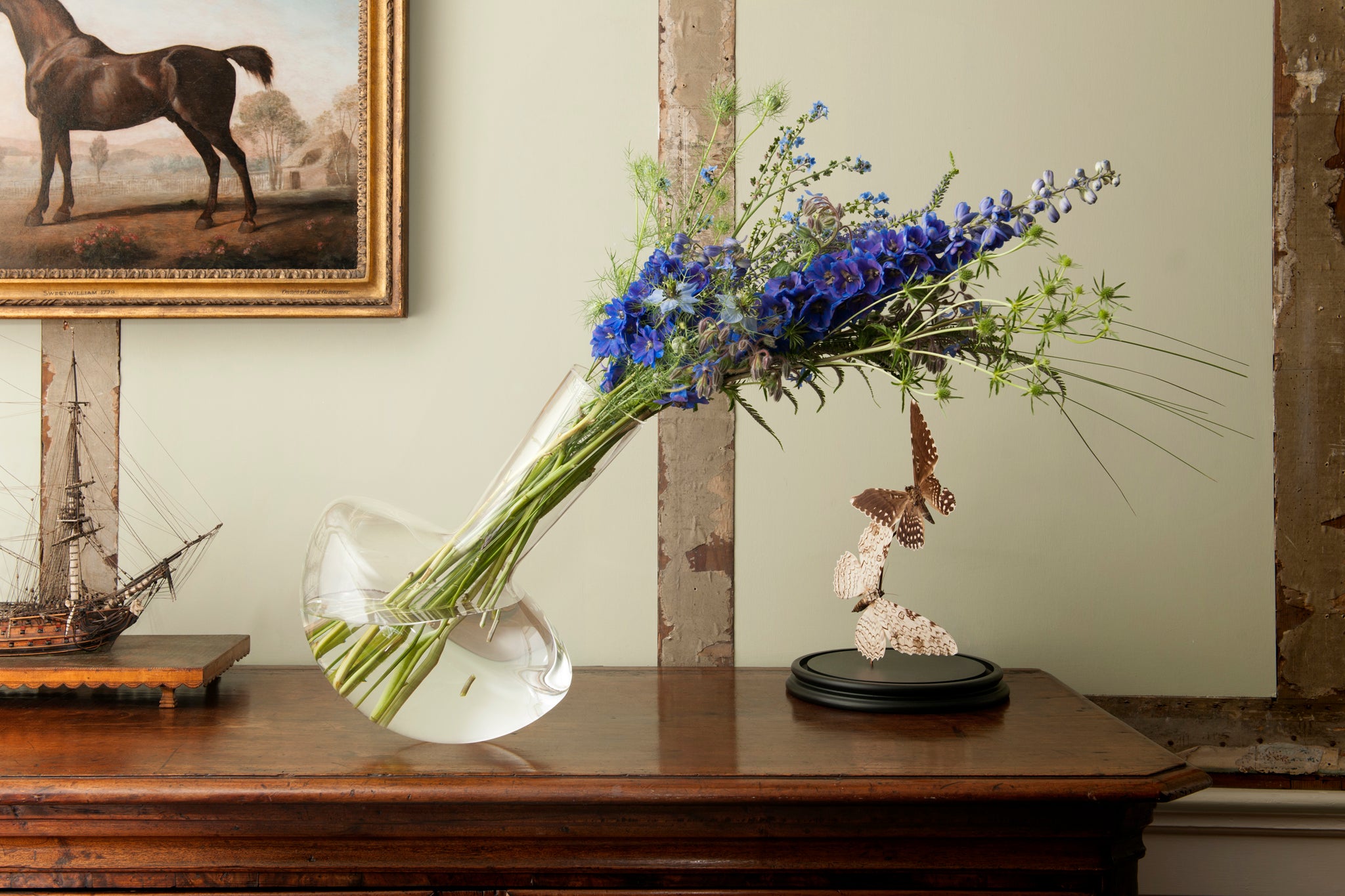 Decorative borosilicate vase with flowers on a wooden chest of draws. A framed picture of a horse hangs in the background. 