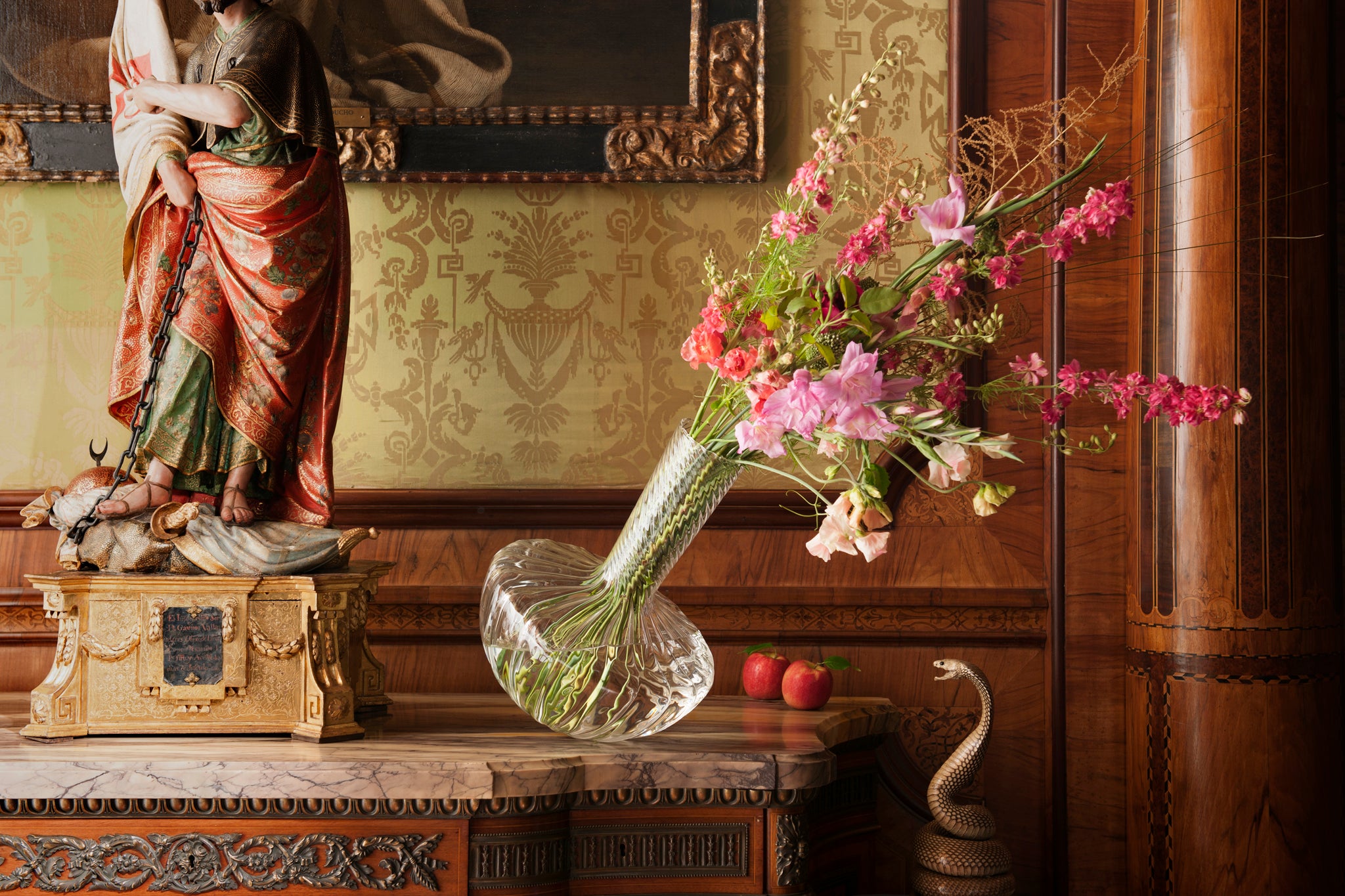 Decorative setting with a statue, borosilicate Gauge vase, and flowers on a marble surface. Two apples decorate the chest of draws and a snake looks on.