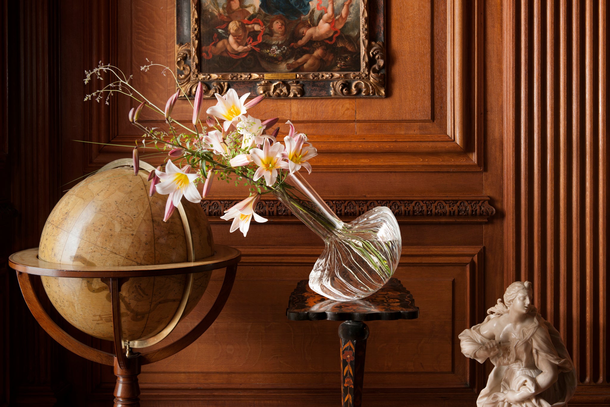 Decorative scene with a borosilicate flower vase leaning dramatically towards a globe, and a classical sculpture against a wooden wall.