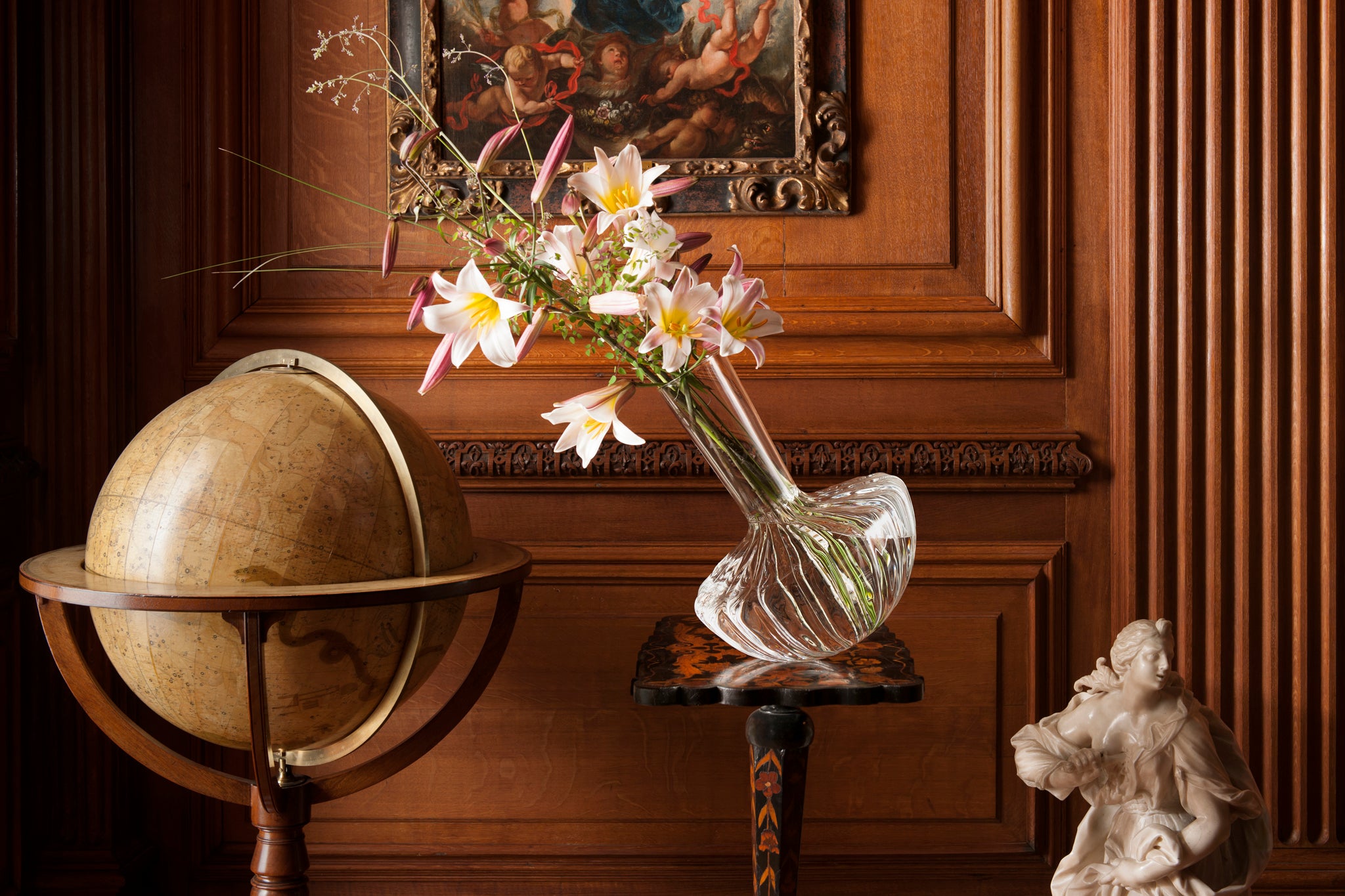 Decorative scene with a borosilicate flower vase leaning towards a globe, and a sculpture against a wooden wall.