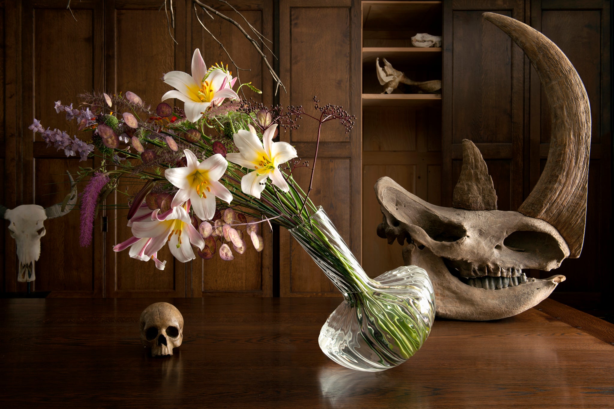 Still life arrangement with flowers in a glass Gauge vase, two skulls on a wooden surface.