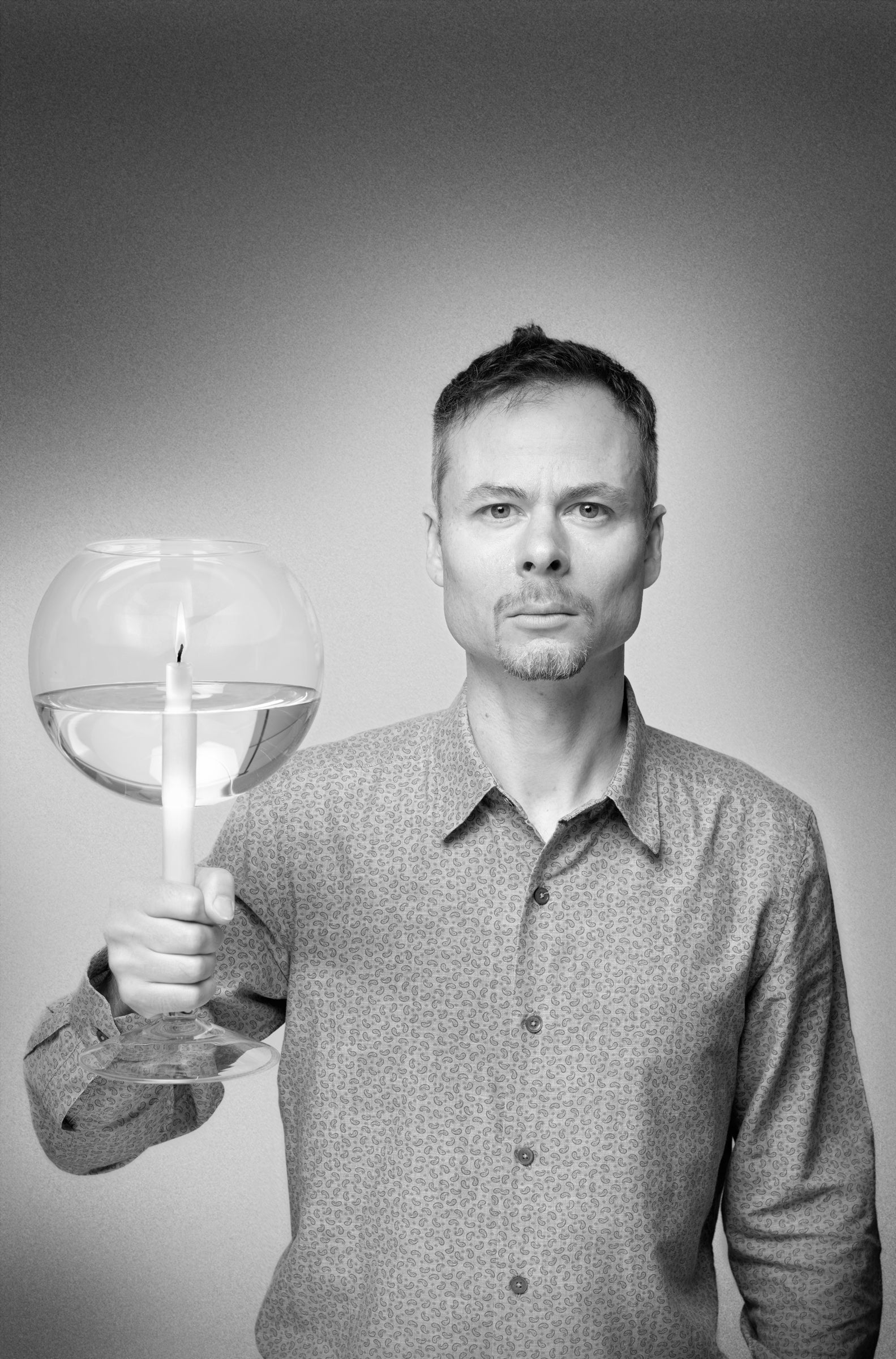 A black and white portrait of a man holding a clear glass bowl filled with water and a lit candle, set against a neutral gray studio background.